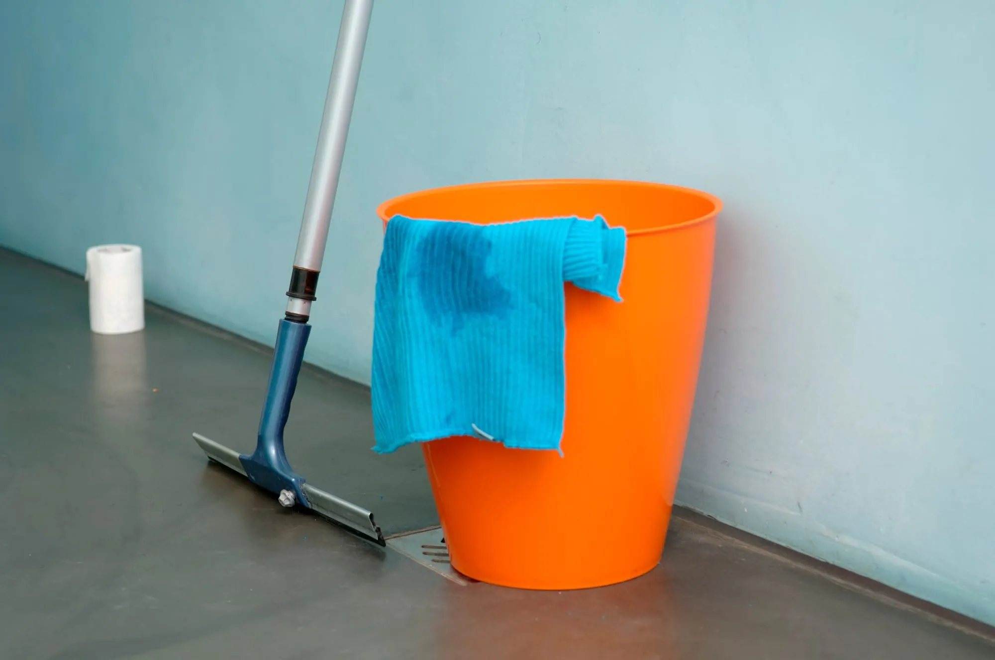 Oranje emmer met een blauwe doek en een trekker met lange steel tegen een lichtblauwe muur